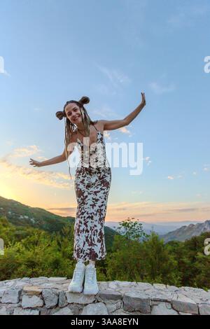 Una donna in un abito che scorre si erge in cima ad una struttura di pietra al tramonto, braccia sollevate con grazia. La sua silhouette contrasta con il cielo vibrante Foto Stock