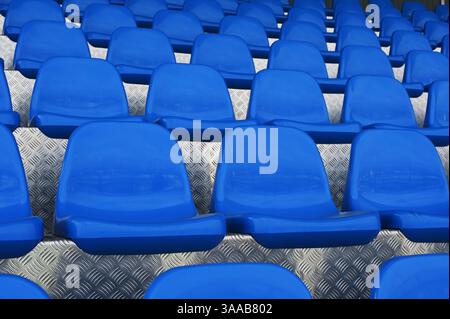 Una fotografia ravvicinata dei sedili vuoti in plastica blu disposti in file simmetriche su un pavimento metallico strutturato. Il colore blu intenso contrasta Foto Stock