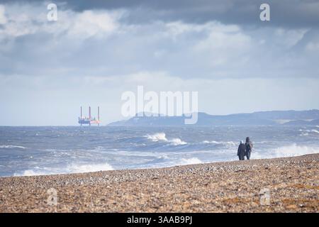 Gente sulla spiaggia di ciottoli in inverno, guardando una lontana piattaforma petrolifera nel Mare del Nord. Blakeney Point, Norfolk, Regno Unito Foto Stock