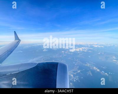 Una vista di un'ala di un aereo che vola sopra un paesaggio di montagna coperto di neve. Catturato dall'alta quota con cieli limpidi e viste sulle montagne. Foto Stock