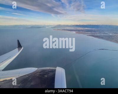 Vista aerea della costa e delle montagne lontane dalla finestra dell'aeroplano con l'ala visibile, catturata durante il volo diurno. Foto Stock