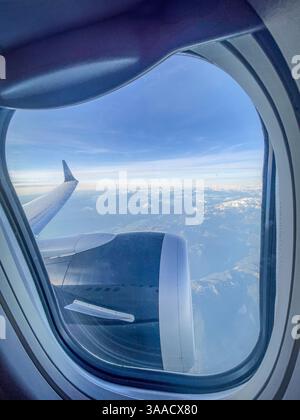 Vista dell'ala e del motore di un aeroplano attraverso il finestrino del passeggero, catturando le cime innevate sullo sfondo durante i viaggi in alta quota. Foto Stock