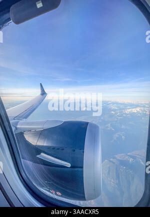 Primo piano di un motore di un aereo e di un'ala visti attraverso la finestra con una vista sulle cime innevate delle montagne e sui cieli blu sullo sfondo. Foto Stock