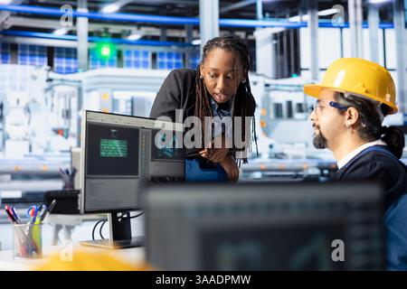 Il supervisore di una fabbrica di fotovoltaici supervisiona il lavoro svolto da esperti per la messa a punto delle impostazioni del sistema sul PC. Il manager afroamericano lavora con l'ingegnere nella configurazione delle linee di assemblaggio degli impianti a pannelli solari Foto Stock