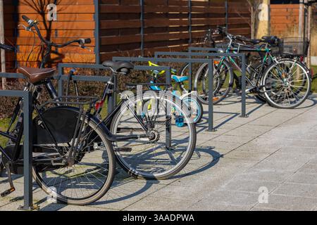 Varie biciclette e una piccola bici di bambini parcheggiate in un portabiciclette in metallo su un marciapiede pavimentato vicino a una recinzione di legno in condizioni di sole Foto Stock