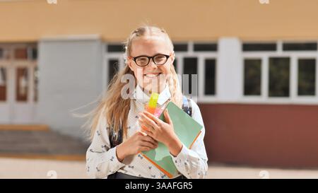 Una studentessa felice sorridente e che tiene libri e governante davanti all'edificio scolastico, pronta per l'apprendimento e l'istruzione Foto Stock