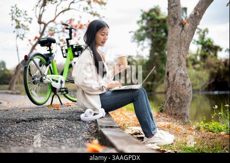 Una donna asiatica sta fissando il portatile mentre tiene un drink in una mano, lavora o studia all'aperto nel parco o in giardino. Rilassatevi nella natura. Foto Stock