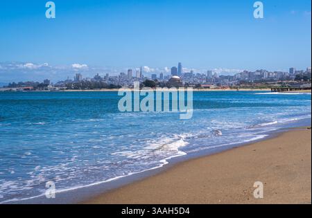 La spiaggia sabbiosa di Golden Gate Beach incontra l'Oceano Pacifico, con lo skyline di San Francisco visibile in lontananza sotto un cielo azzurro Foto Stock