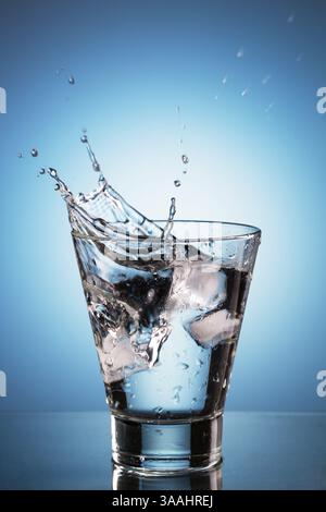 Il cubo di ghiaccio schizzi in un glassl d'acqua. Sfondo blu Foto Stock