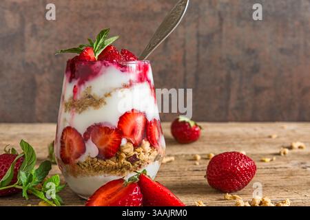 Parfait di yogurt alla fragola con muesli e menta fresca in un bicchiere con cucchiaio su un tavolo rustico in legno con frutti di bosco interi. Colazione salutare. Cibo gustoso Foto Stock