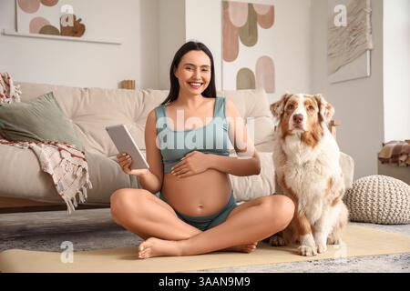 Giovane felice donna incinta sportiva con tablet e carino cane pastore australiano seduto sul tappetino da yoga a casa Foto Stock