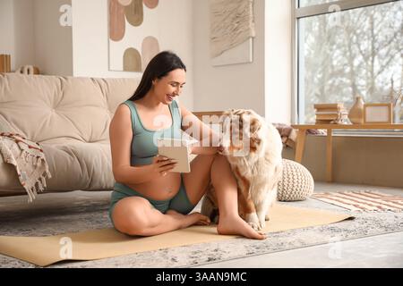 Giovane felice donna incinta sportiva con tablet e carino cane pastore australiano seduto sul tappetino da yoga a casa Foto Stock