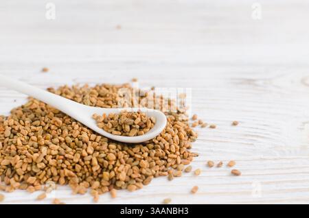 Un mucchio di fieno greco secco o di shambhala con un piccolo cucchiaio da tè su un tavolo di legno bianco. Pianta medicinale dal Medio Oriente. Concetto di medicina alternativa. SEL Foto Stock