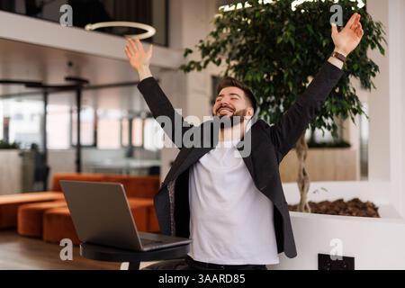 Eccitato gridare giovane uomo guardando lo schermo del suo computer e trionfare con le mani sollevate Foto Stock