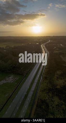 Un video registrato durante l'ora d'oro, che mostra una strada tortuosa attraverso un paesaggio rurale, campi con riflessi d'acqua e edifici lontani, il tutto sotto Foto Stock