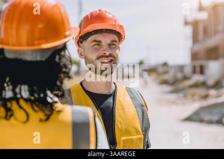 Il muratore felice sorriso, ingegnere caucasico maschio in piedi che indossa giubbotto di sicurezza e casco nel cantiere. Giovane e cordiale architetto watchi Foto Stock