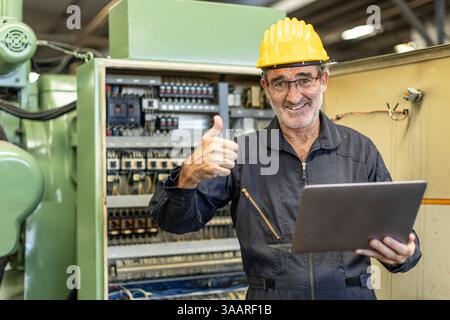 Senior Professional Electrician ingegnere supervisore felice sorridente lavoro in macchine per l'industria pesante cavo elettrico con computer portatile. Foto Stock