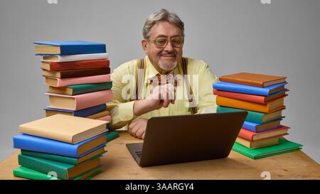 Professore anziano circondato da libri colorati, che lavora su un computer portatile con una matita in mano su sfondo grigio studio Foto Stock