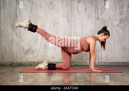Atleta in gravidanza che si esercita con pesi alla caviglia, esegue un calcio alla schiena, un'attività prolungata al ginocchio sul tappetino da yoga Foto Stock
