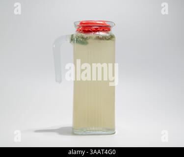 Caraffa in vetro trasparente con limonata e foglie di menta fresca chiusa con coperchio rosso su fondo liscio. Foto Stock