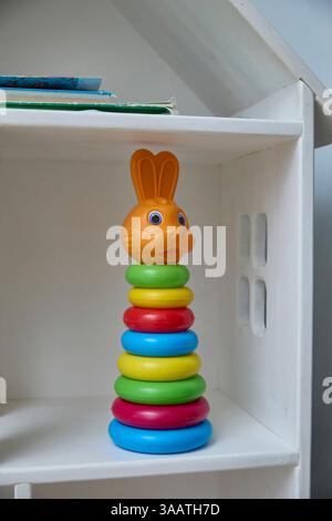 Allegro giocattolo impilabile per bambini con anelli colorati e una vivace testa a coniglietto posizionata ordinatamente su uno scaffale bianco, probabilmente utilizzato per l'intrattenimento o per i primi anni Foto Stock