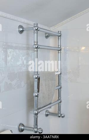 Portasciugamani in cromo fissato a una parete del bagno con piastrelle bianche floreali, con un asciugamano beige appeso in modo ordinato, che aggiunge eleganza e funzionalità alla spac Foto Stock