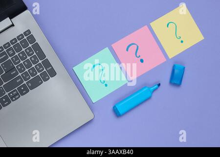 Laptop, appunti di carta con punti interrogativi disegnati e pennarello su sfondo viola. Vista dall'alto Foto Stock