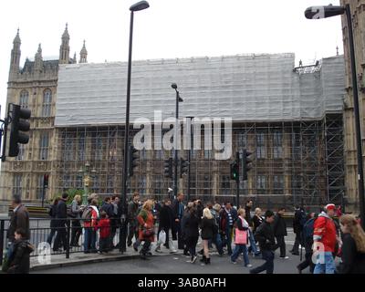 Un'animata strada con una folla di pedoni che attraversano di fronte a un grande edificio in fase di ristrutturazione, coperto da impalcature e lenzuola protettive Foto Stock