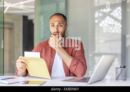 Ritratto di un uomo turbato in ufficio, il dipendente ha ricevuto una lettera con notizie negative e tristi per posta. Uomo d'affari che tiene in mano una busta con notifica e guarda la macchina fotografica. Foto Stock