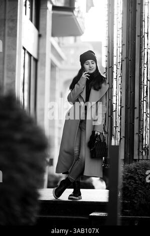 Vestita con un cappotto e un berretto eleganti, una giovane donna si appoggia in modo informale contro una ringhiera, assaporando una telefonata in uno sfondo urbano pieno di gemme Foto Stock