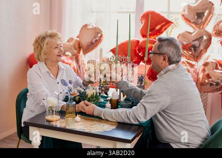 Una romantica coppia anziana che festeggia San Valentino o anniversario a casa, brindando con i bicchieri e tenendo per mano un tavolo decorato con il cuore Foto Stock