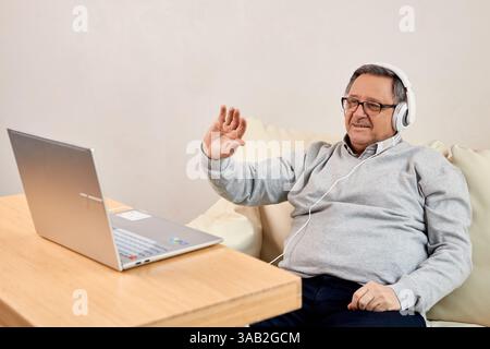 Uomo anziano con le cuffie che sventolano durante una videochiamata sul proprio computer portatile a casa. Ideale per la comunicazione online, la tecnologia per anziani e il collegamento remoto Foto Stock