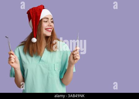 Ritratto di una giovane dentista femminile nel cappello di Babbo Natale con attrezzi su sfondo viola Foto Stock