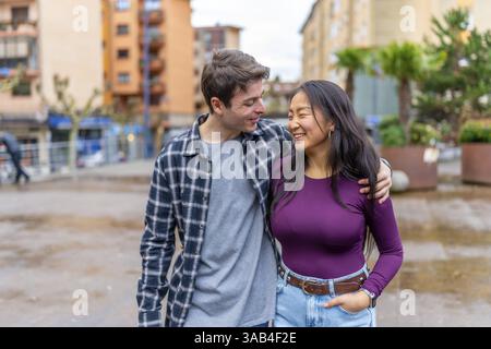 Una coppia multietnica gioiosa condivide un momento felice all'aperto, abbracciandosi e sorridendo l'un l'altro, circondati da edifici urbani e vegetazione Foto Stock