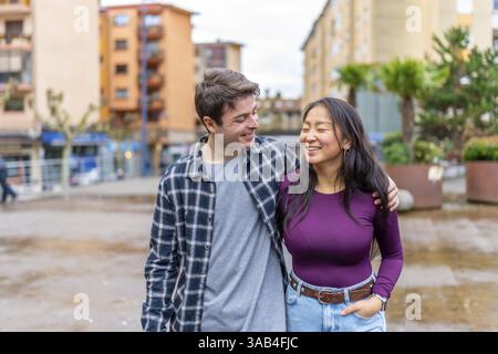 Una coppia multietnica cammina braccio nel braccio, condividendo un momento di gioia in un ambiente urbano. La scena cattura un mix di culture e felicità Foto Stock