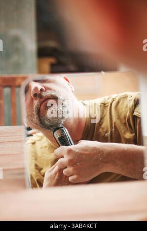 Primo piano di un uomo che rifinisce la barba all'aperto con un rasoio elettrico e un sistema di cura fai da te Foto Stock