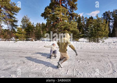 Una coppia matura ama divertirsi in inverno, mentre un uomo tira la moglie su una slitta attraverso il terreno innevato in un ambiente all'aperto vicino agli alberi mentre si dedicano ad attività ricreative. Foto Stock