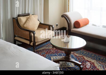 Camera d'hotel con vista su sedia, divano e parte di un letto singolo ricoperto di coperta bianca. Foto Stock