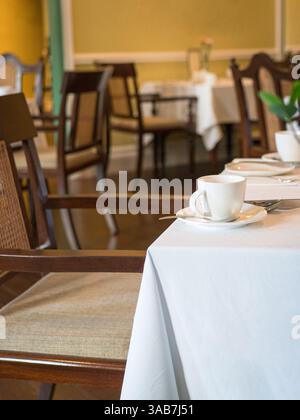 Interno di un ristorante. Primo piano su una tazza bianca e un piattino sul tavolo ricoperto di panno bianco. Foto Stock