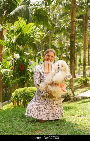 Donna sorridente inginocchiata su un'erba lussureggiante tenendo in mano il suo piccolo cane bianco vestito con un grazioso abito rosa, circondato da vivaci piante tropicali in un sole Foto Stock