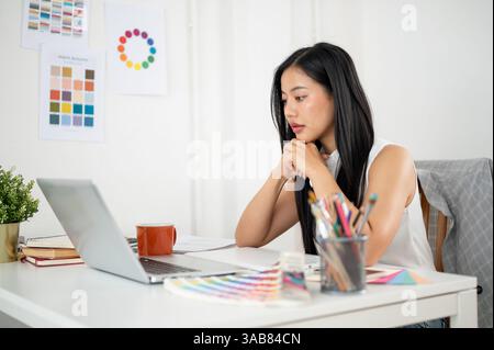 Una donna asiatica sta mettendo entrambe le mani sotto il mento e fissando con attenzione lo schermo del laptop sul tavolo. Interior Designer o Graphic Designer che lavorano i Foto Stock