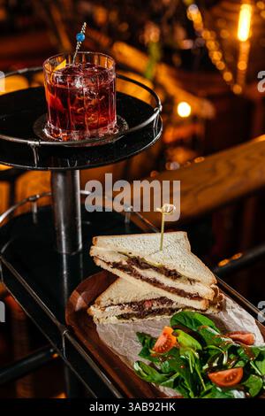 Un panino da club gourmet pieno di teneri guance di vitello, servito insieme a un'insalata di pomodori e spinaci freschi. Accompagnato da un classico cocktail negroni Foto Stock