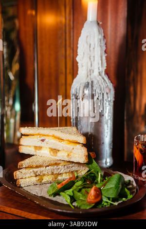 Un club sandwich deliziosamente impilato, ripieno di formaggio fuso e cipolle caramellate, servito con un contorno di insalata fresca. Un ambiente rustico a lume di candela e. Foto Stock