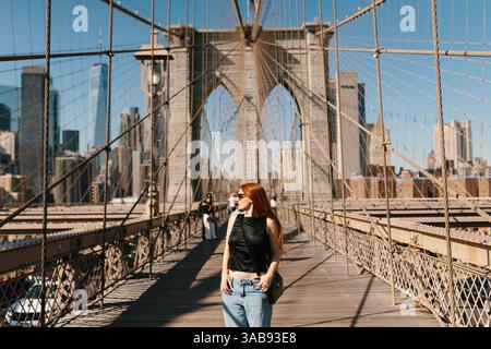Una donna con i capelli rossi attraversa il ponte di Brooklyn, crogiolandosi al sole. L'iconico ponte intricati cavi e lo skyline di New York sono un vero e proprio punto di riferimento Foto Stock