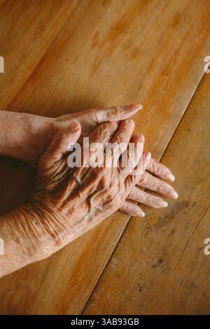 Primo piano di mani anziane che poggiano delicatamente su un tavolo di legno, catturando la consistenza e il calore dell'invecchiamento. L'illuminazione da studio migliora i dettagli e l'ombra Foto Stock