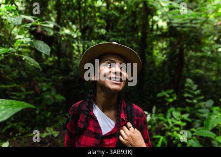 Una donna ecuadoriana, vestita con una camicia a quadri rossa e dotata di uno zaino da trekking, sorride con gioia mentre esplora la fitta e verde pioggia di Choco Andino Foto Stock