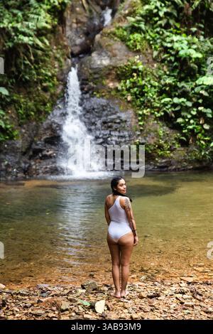 La vista posteriore di una giovane donna ecuadoriana con un costume da bagno bianco si erge ammirando una suggestiva cascata nella lussureggiante foresta pluviale Choco Andino dell'Ecuador Foto Stock