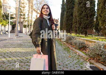 Una donna gioiosa con un cappotto verde porta borse della spesa e un piccolo chihuahua mentre cammina lungo una tranquilla strada della città costeggiata da alberi, godendosi un sole Foto Stock