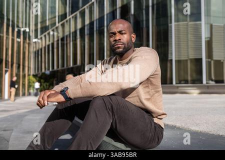 Un uomo cubano in un abbigliamento informale si siede all'esterno di un moderno edificio in vetro, incarnando un'atmosfera urbana rilassata. La luce del sole mette in risalto le linee architettoniche e le sue Foto Stock
