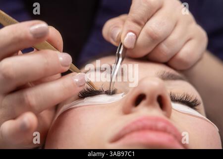 Primo piano di un estetista che applica prolunghe per ciglia con pinzette. L'attenzione è rivolta al lavoro delicato, evidenziando l'esperienza e la cura del salone. Foto Stock